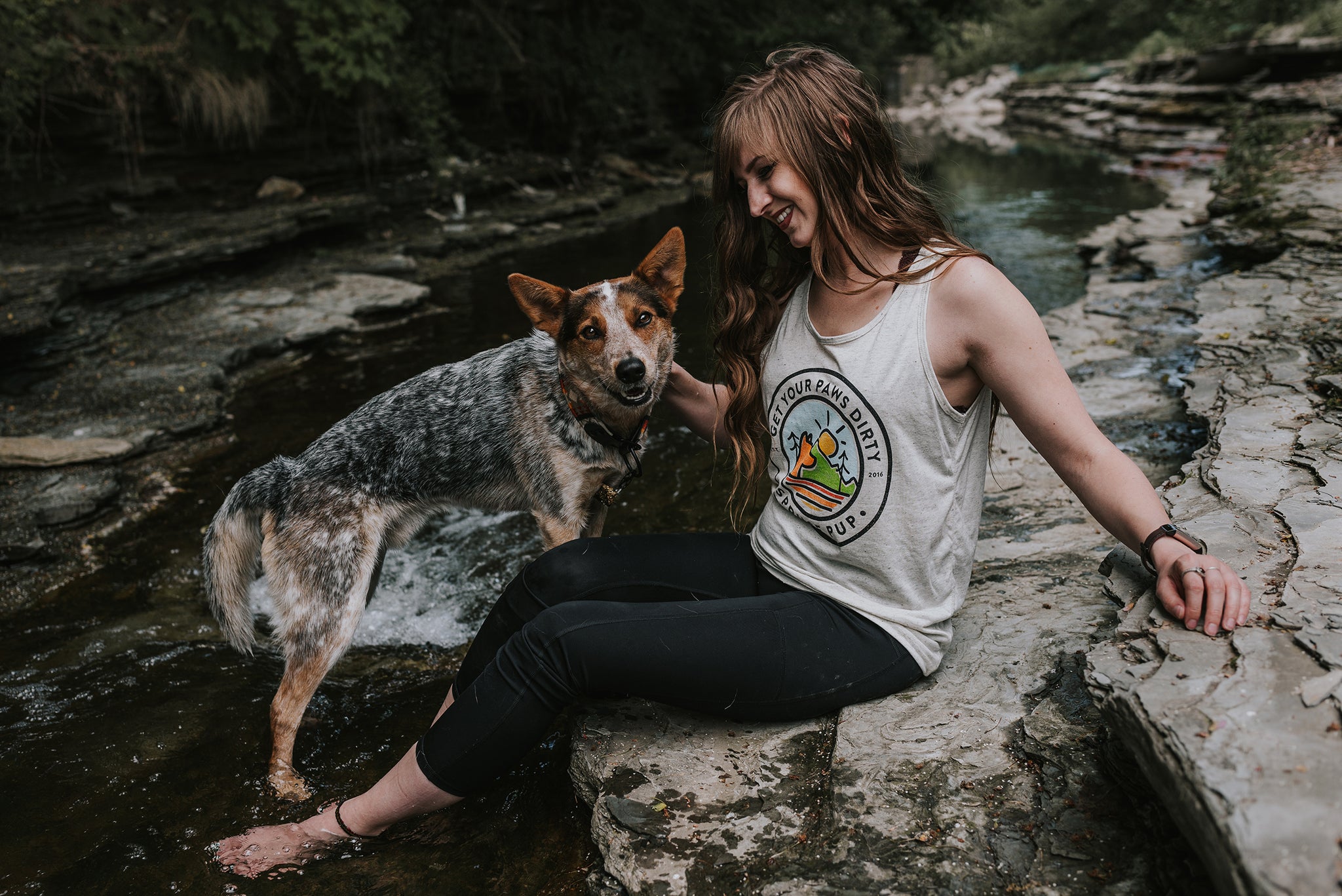Woman wearing Get Your Paws Dirty tank top, sitting by a stream with her pup