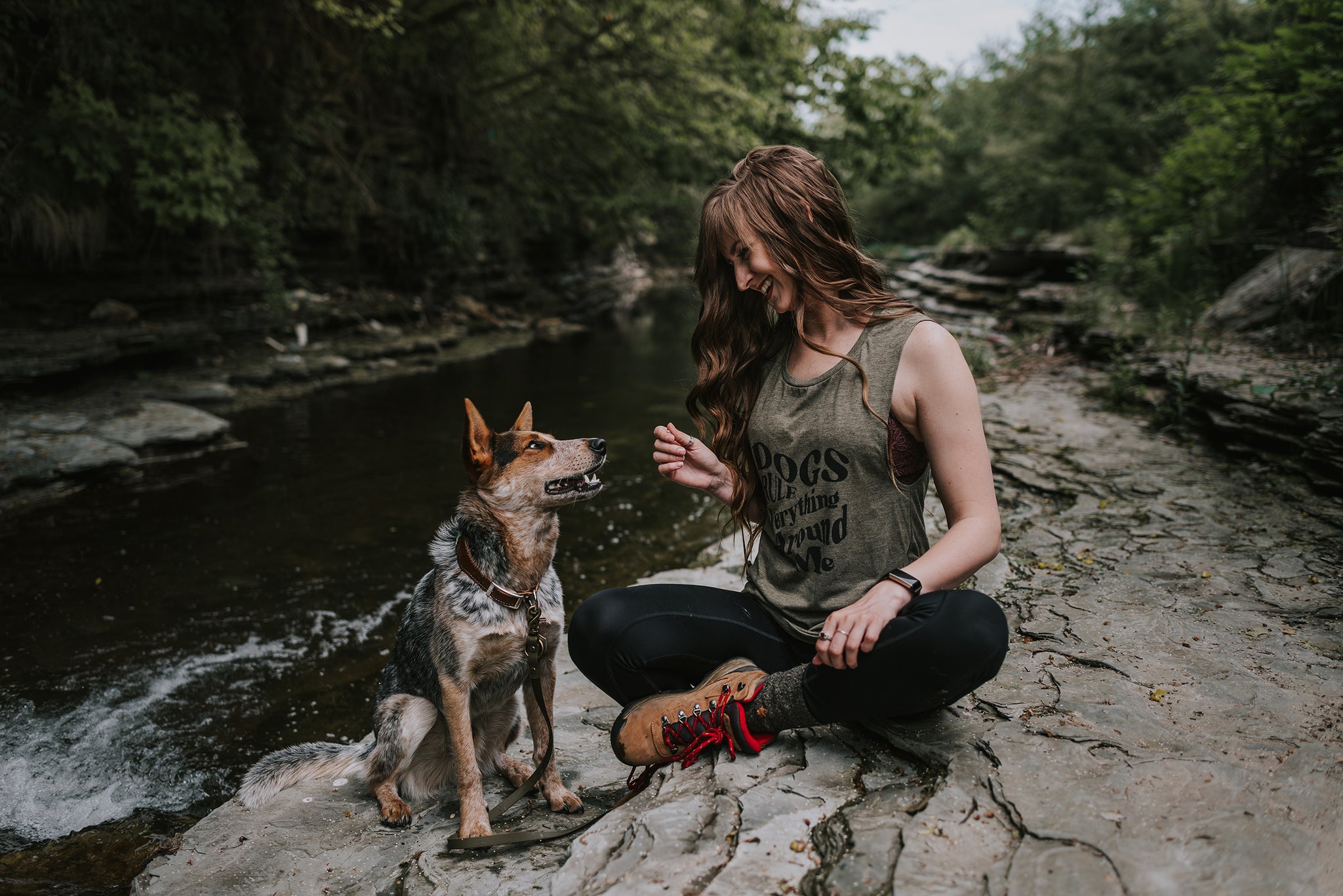 Woman wearing 'Dogs Rule Everything Around Me' tank top sitting next to her dog by a river.